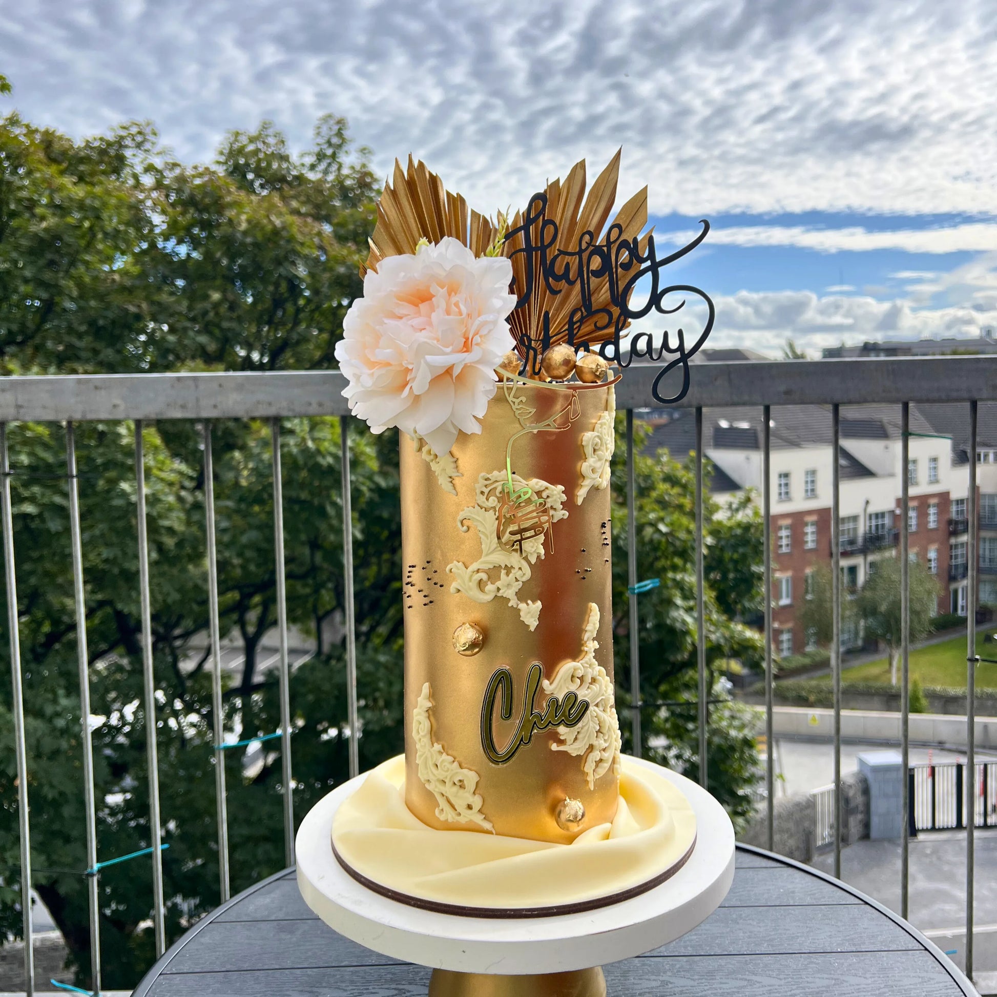 Gold birthday cake with floral decorations on a balcony with a view of trees and buildings.