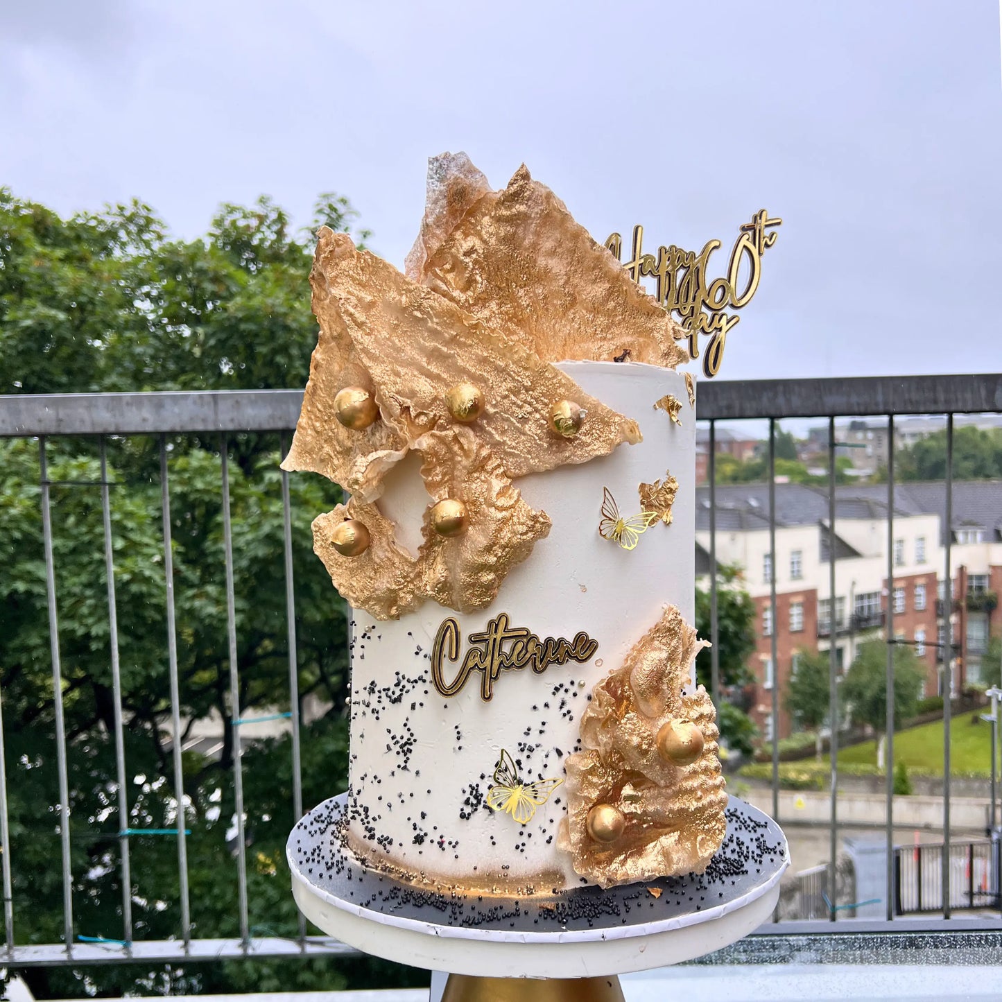 Decorative cake with gold leaf and 'Catherine' on a stand outdoors.