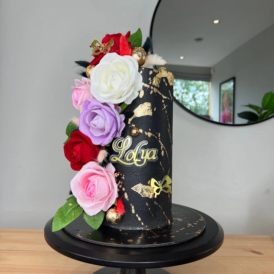 A decorated cake with black buttercream and gold accents, topped with a variety of artificial flowers and the name 'Lola' written on it.