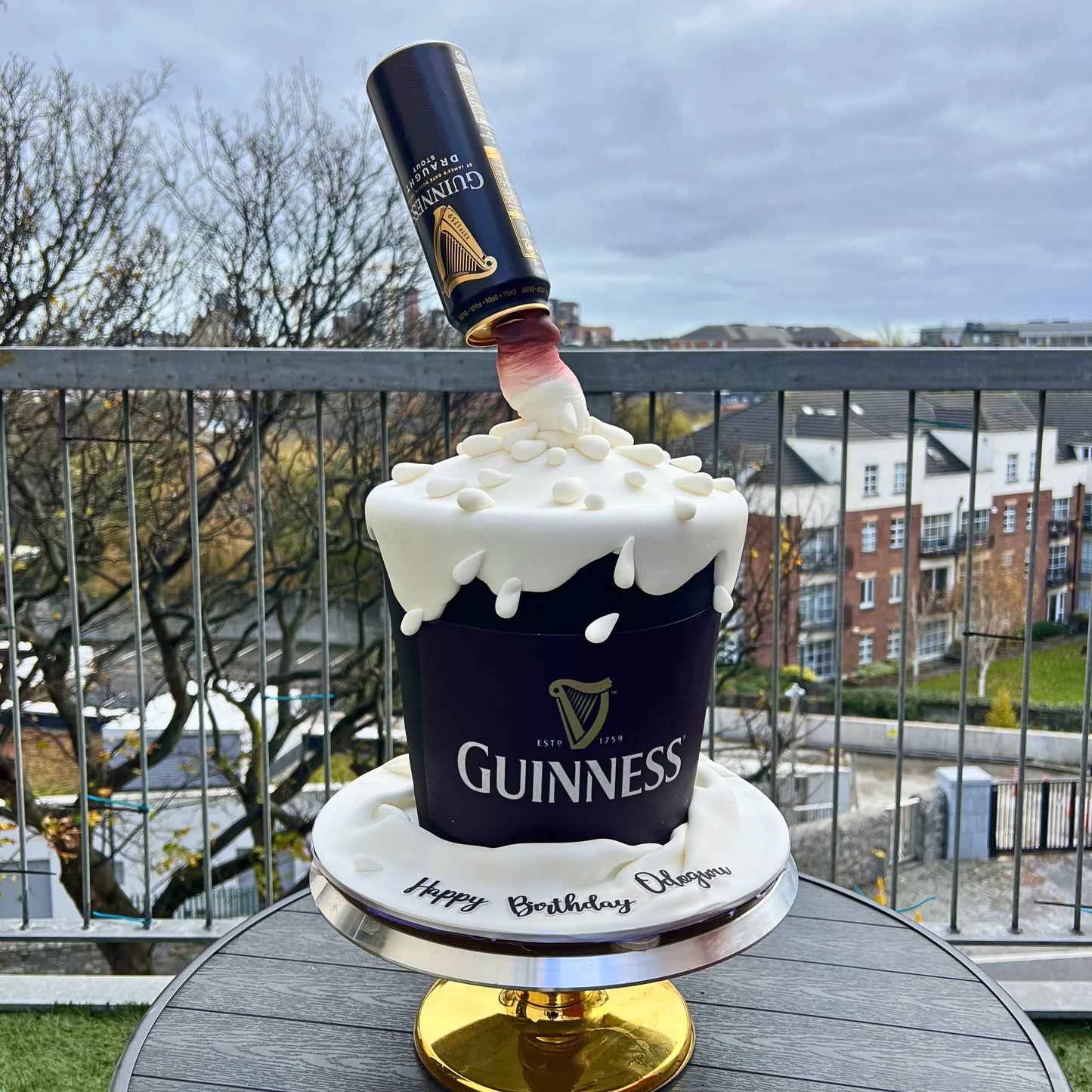 Guinness bottle cake with a bottle on top, outdoors with a view of buildings.