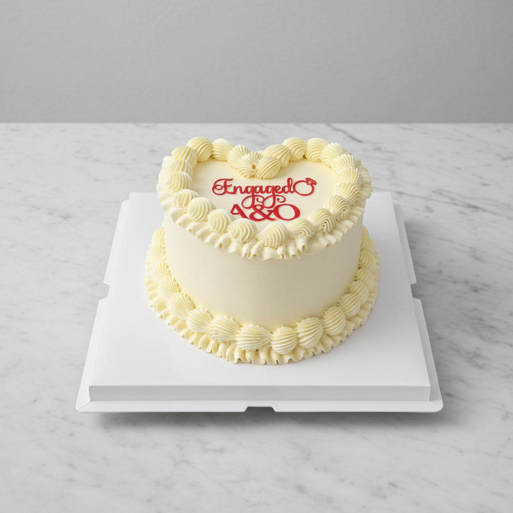 A heart shaped white frosted birthday cake with piped cream borders and red icing text reading 'Engaged 28' on top, displayed on a white square cake box with an outdoor city street and apartment buildings in the background.