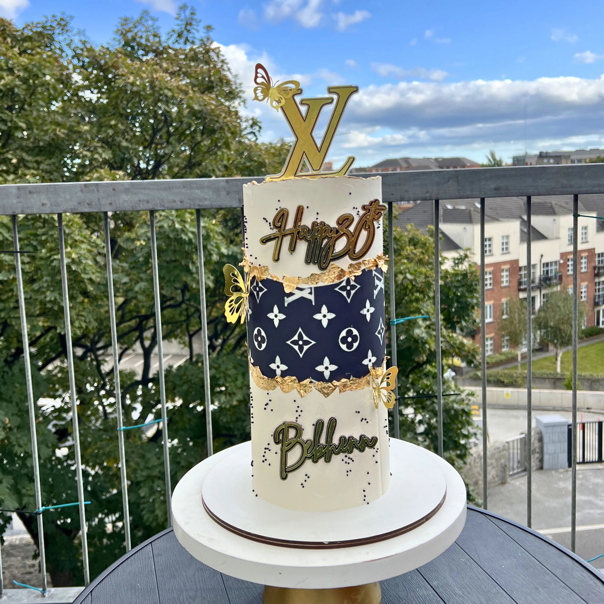 Decorative cake with Louis Vuitton branding on a balcony with a view of trees and buildings.