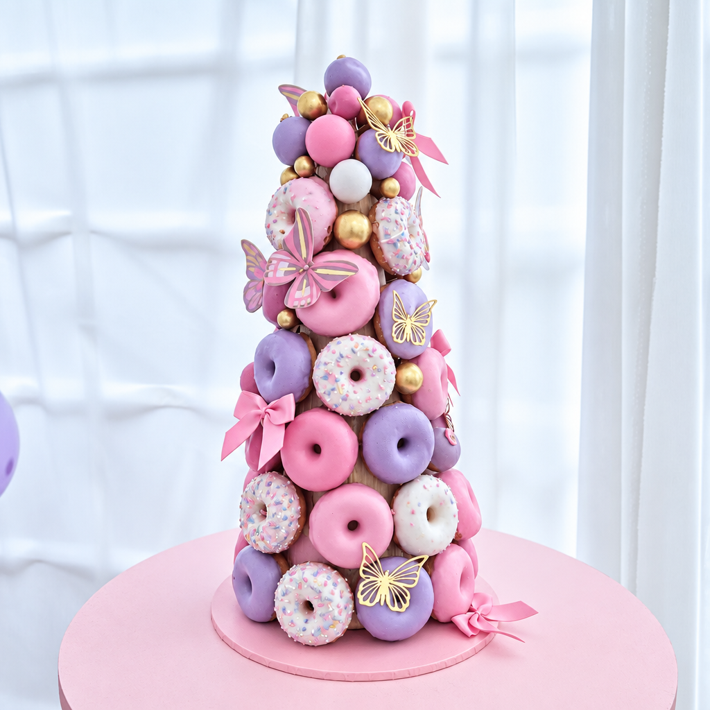A decorative cake shaped like a tower of donuts in shades of pink, mauve, and burgundy, decorated with pink bows, gold ball accents, and butterfly decorations, displayed on a round pink stand in front of a white curtain background.
