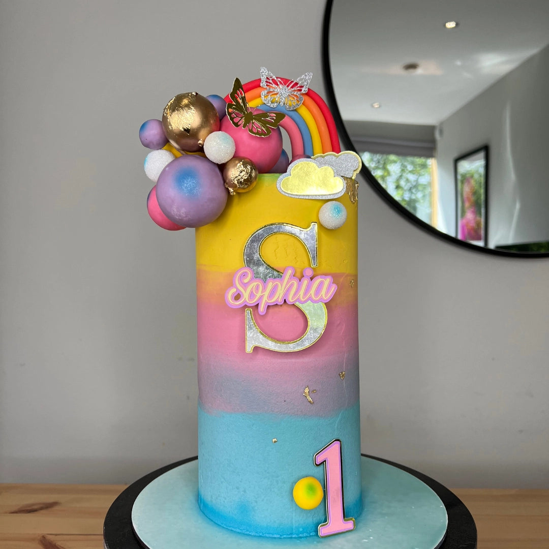 A colorful, themed birthday cake with rainbow layers and decorative elements, including a name in cursive script and age