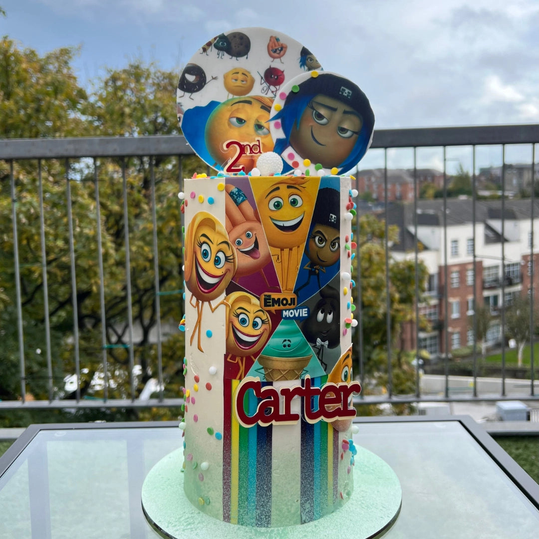 A colorful birthday cake with 'EMOJI MOVIE THEMED CAKE' written on it and decorated with cartoon characters from the Emoji Movie.