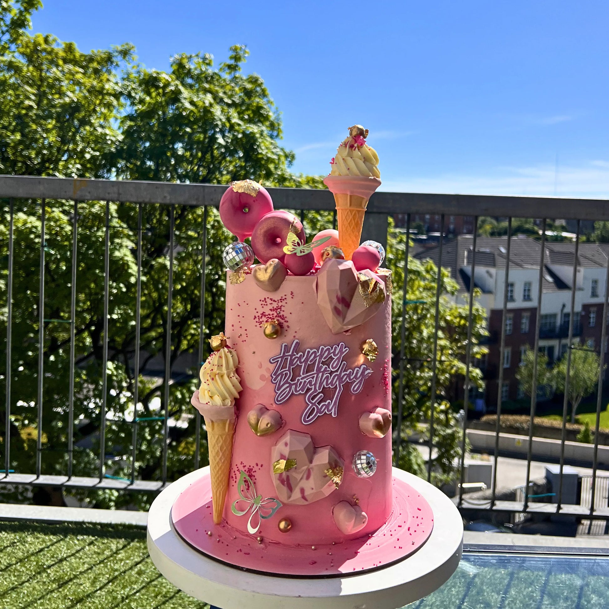 Pink birthday cake with ice cream cones and 'Happy Birthday' text on a balcony with trees and buildings in the background.