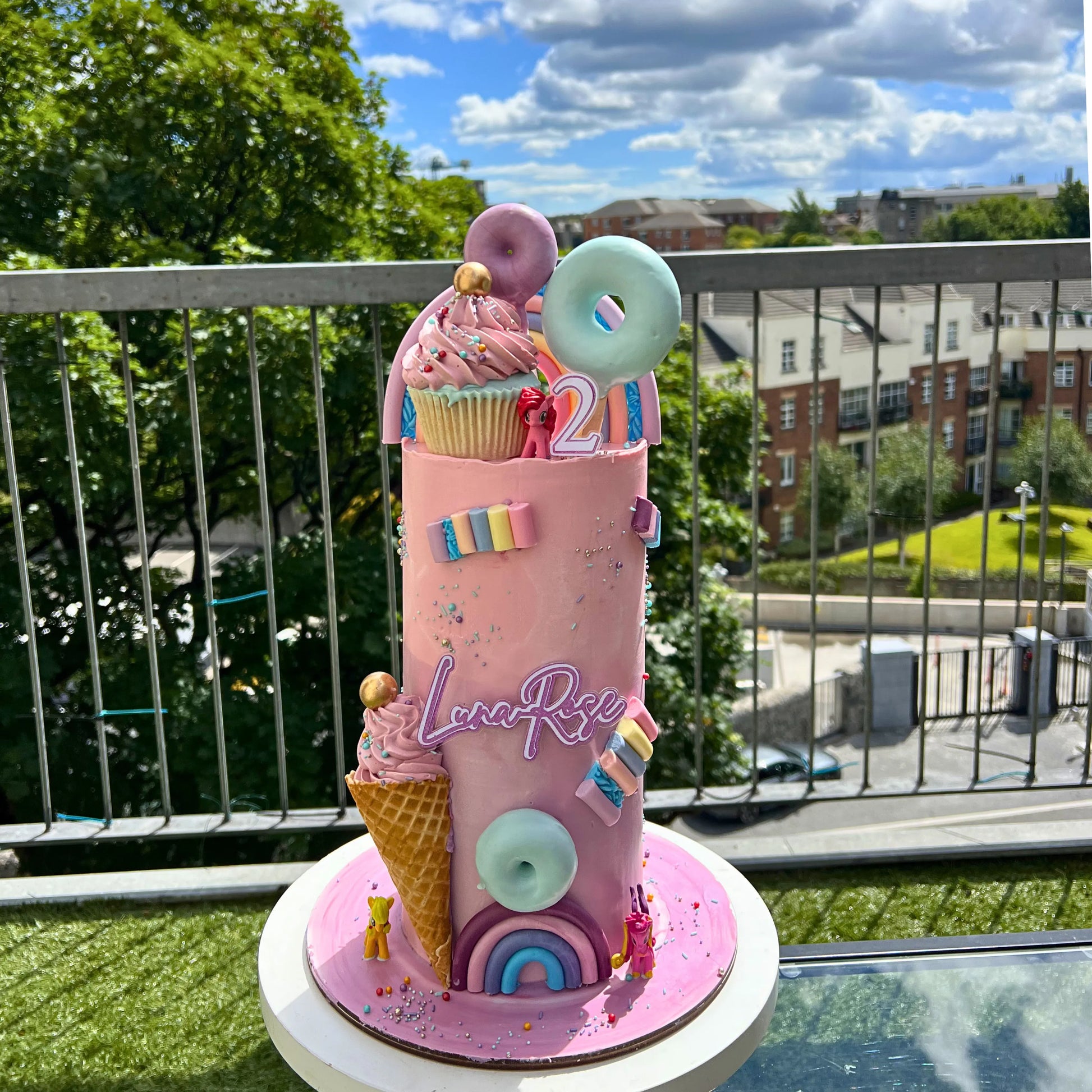 Colorful birthday cake with ice cream cone and rainbow decorations on a balcony with greenery and buildings in the background.