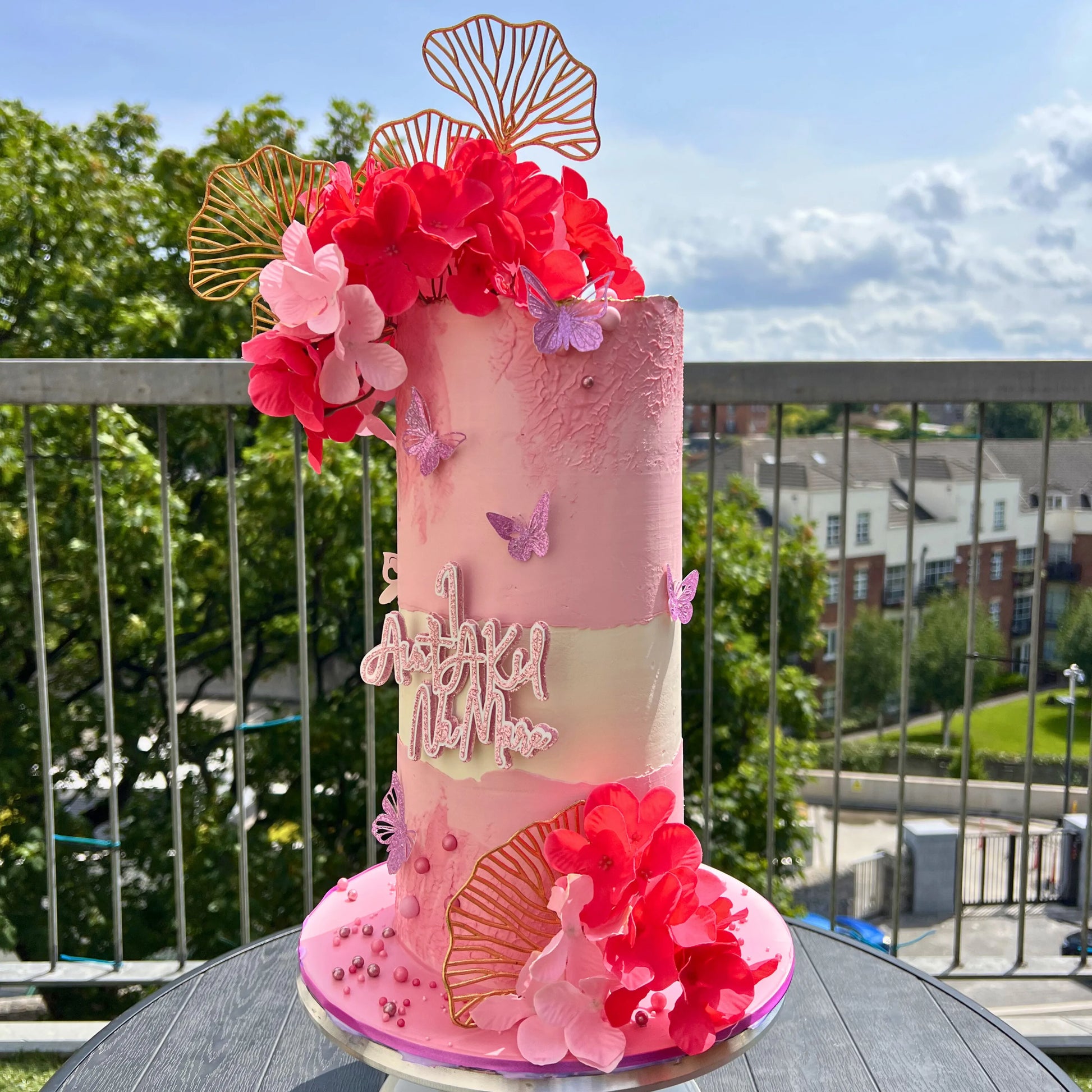 Pink birthday cake with floral decorations on a table outdoors.