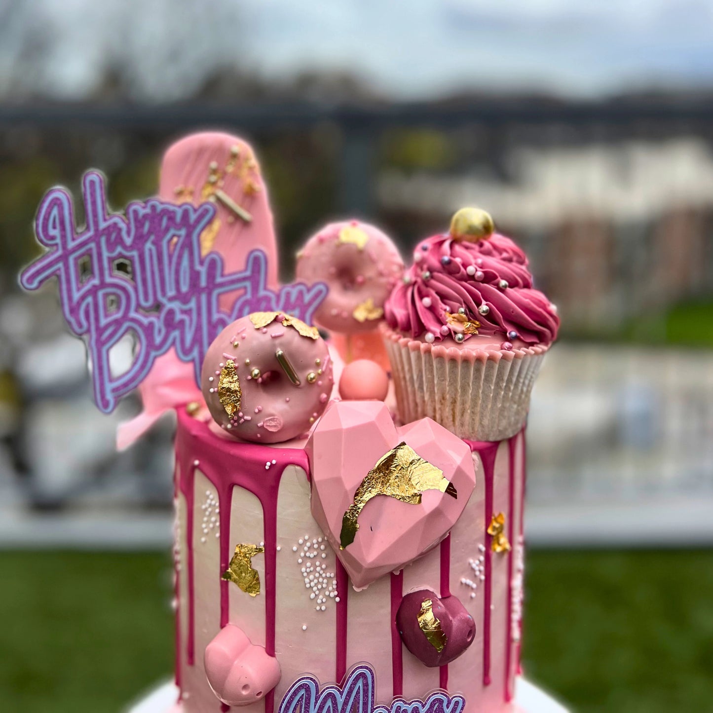 Pink birthday cake with decorative elements and 'Happy Birthday' topper outdoors.
