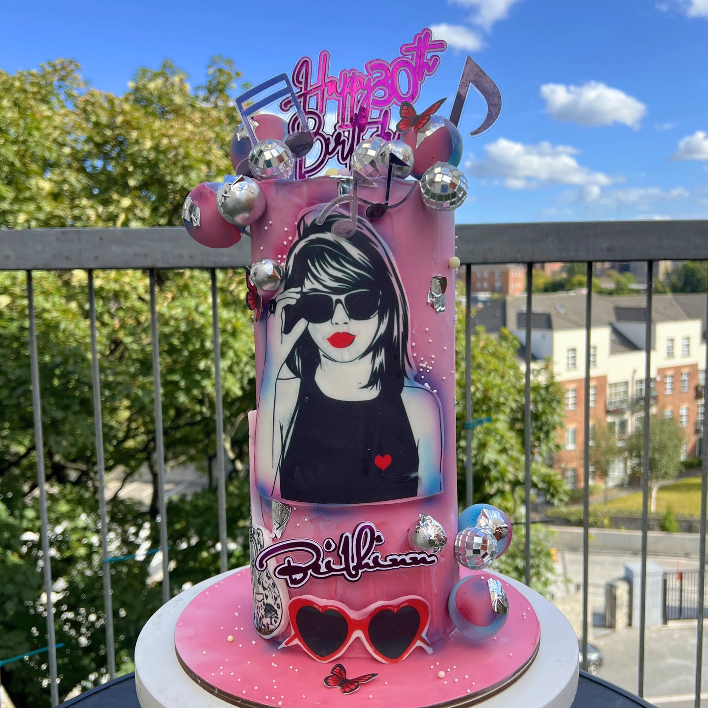 Pink taylor swift birthday cake with decorative elements and a portrait on a balcony with trees and buildings in the background.