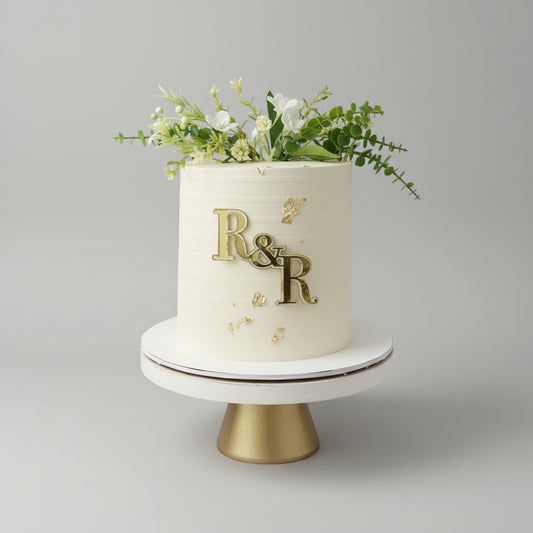 Tall single-tier wedding cake with textured ivory buttercream, fresh flowers, gold leaf and gold monogram plaque, Dublin
