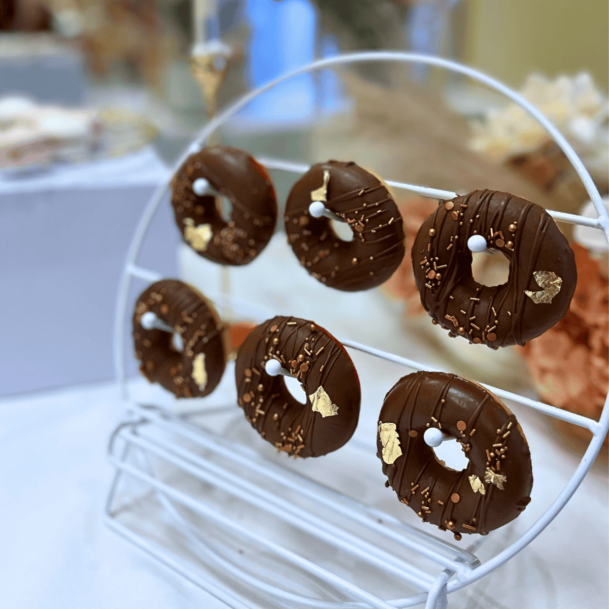 Six chocolate-glazed donuts with gold leaf flakes and sprinkles displayed on a white wire stand in a softly blurred indoor setting.