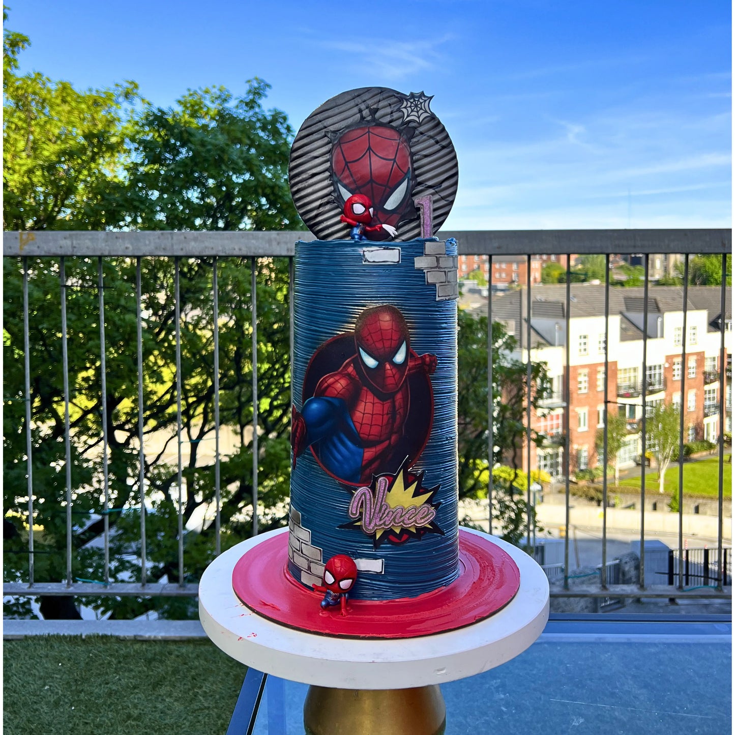 Spider-Man themed birthday cake on a balcony with trees and buildings in the background