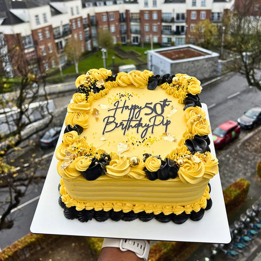 Yellow birthday cake with black text and decorations on a white plate, set against an urban background.