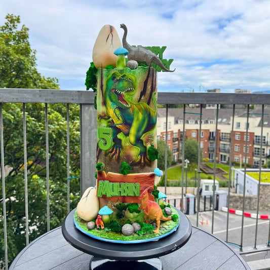 A custom dinosaur-themed birthday cake decorated with a green T-Rex, a small Triceratops, dinosaur eggs, mushrooms, and the name 'Muhsin' on a balcony with a city view in the background.