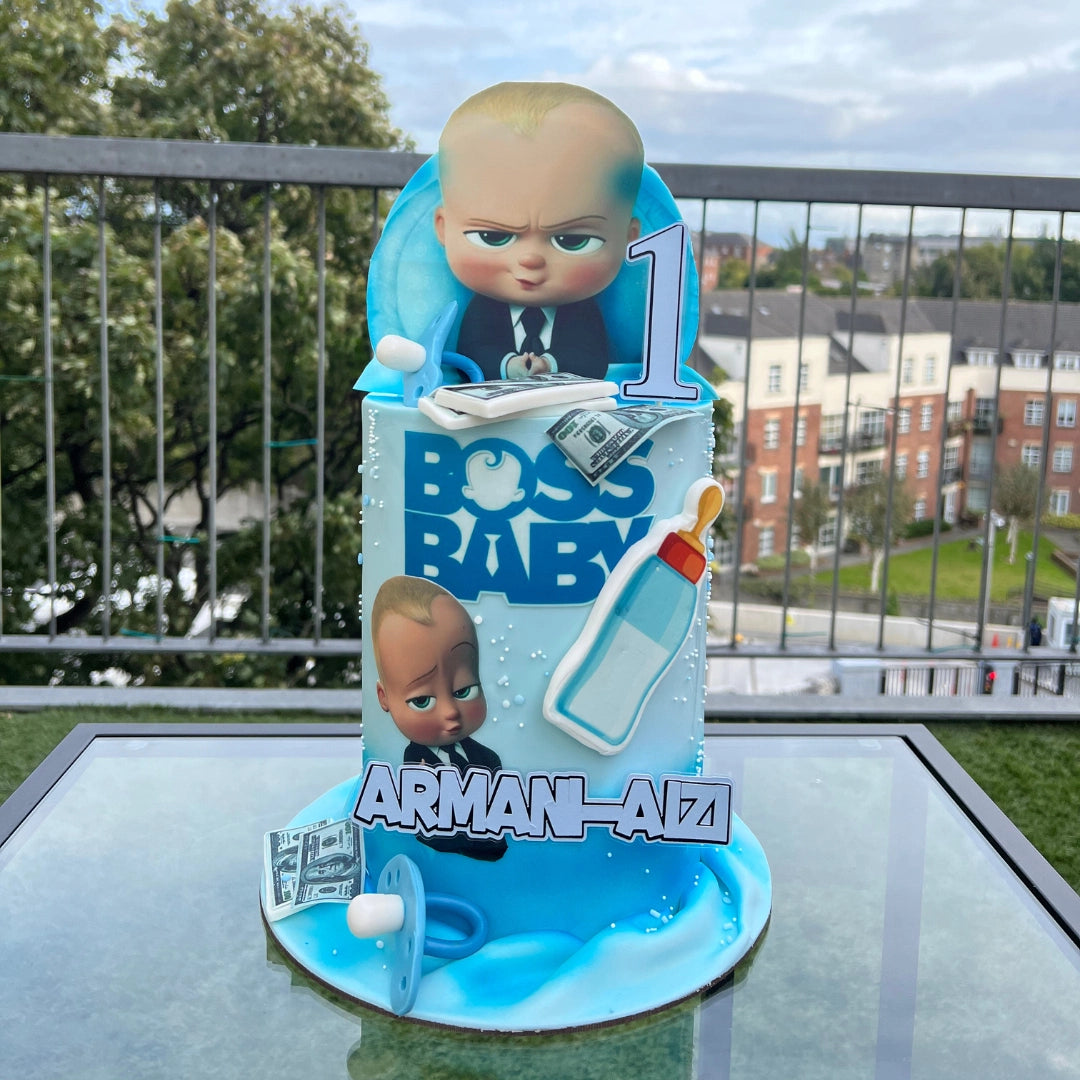 A decorated cake with 'BOSS BABY' theme, featuring cartoon character figurines, blue color scheme, and the name 'ARAM-ALI' printed on the front.