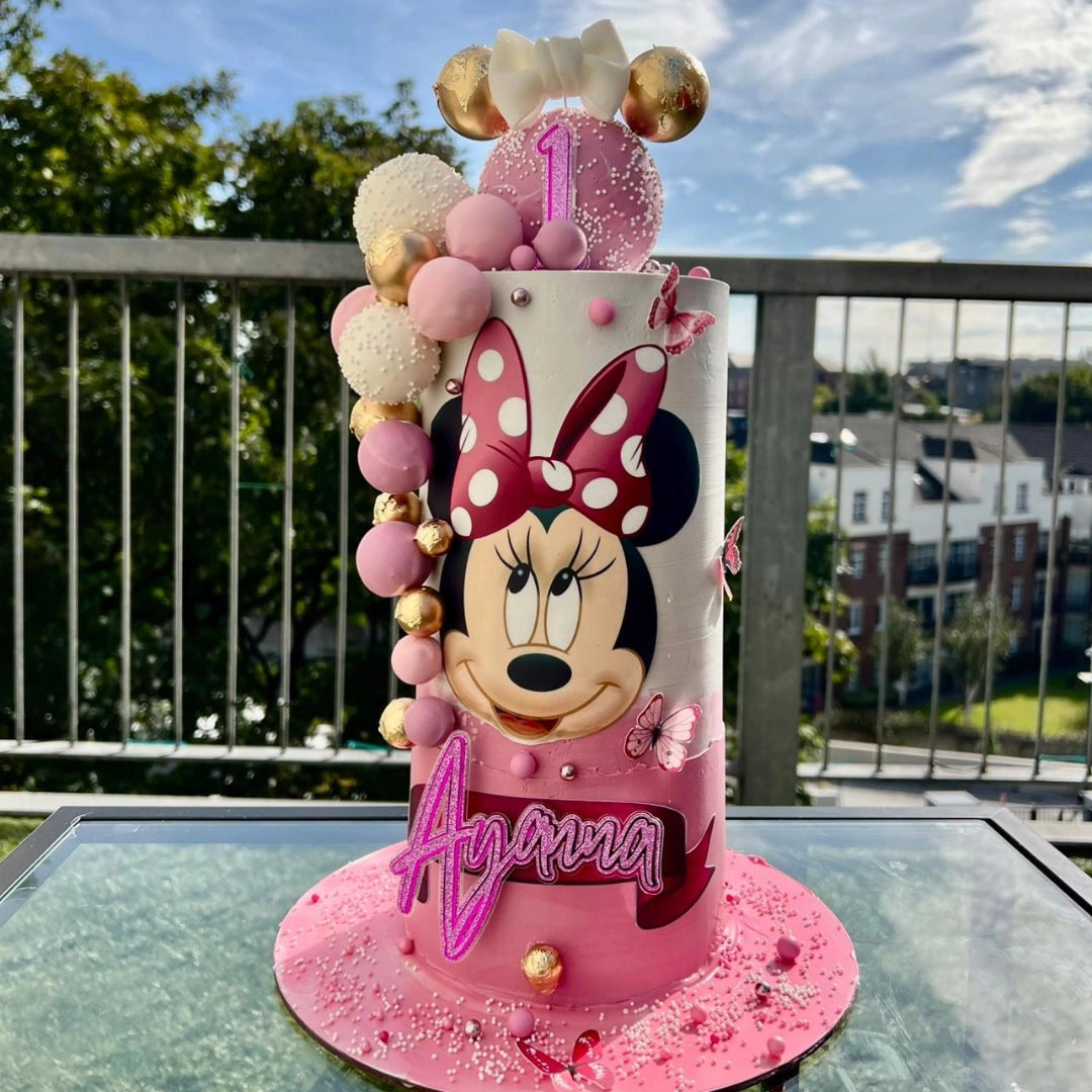 A Minnie Mouse themed birthday cake with pink and gold decorations, topped with a character figurine, and personalized with the name 'Ariana'.