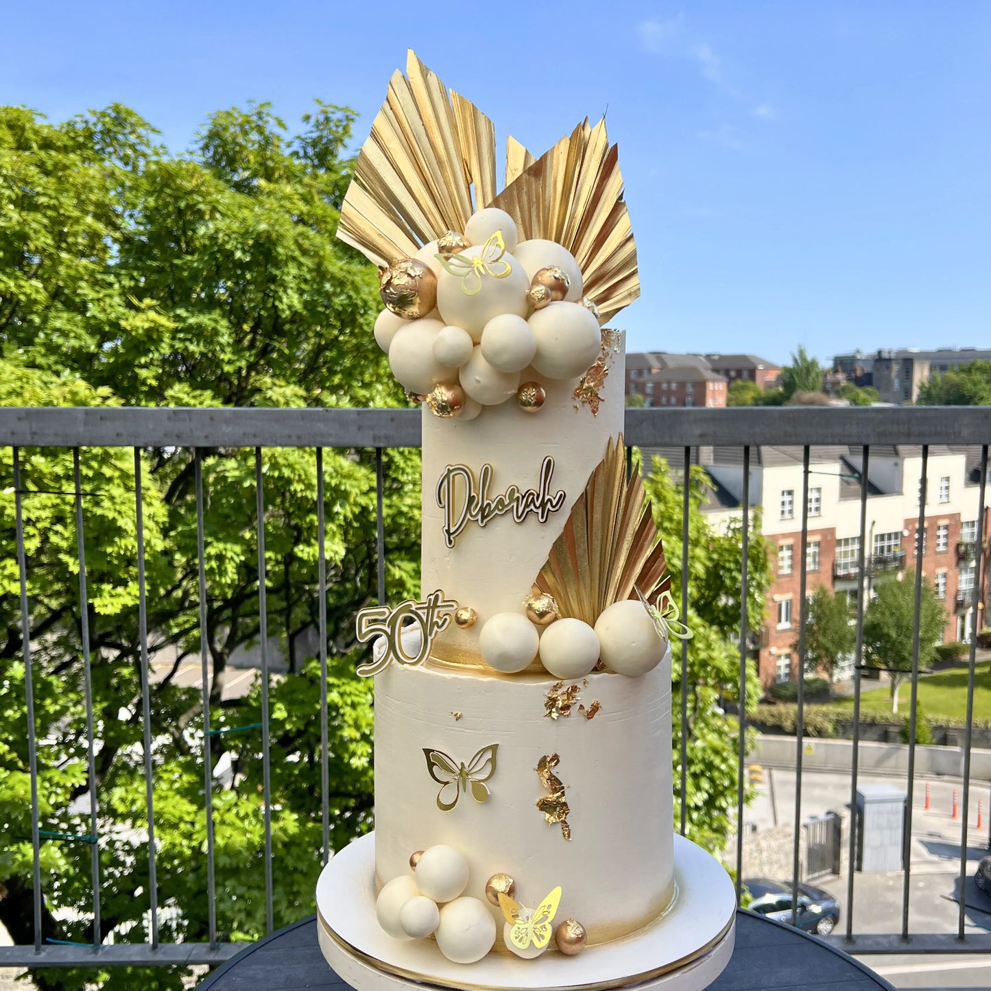 Decorative cake with gold and white elements on a balcony with greenery and buildings in the background