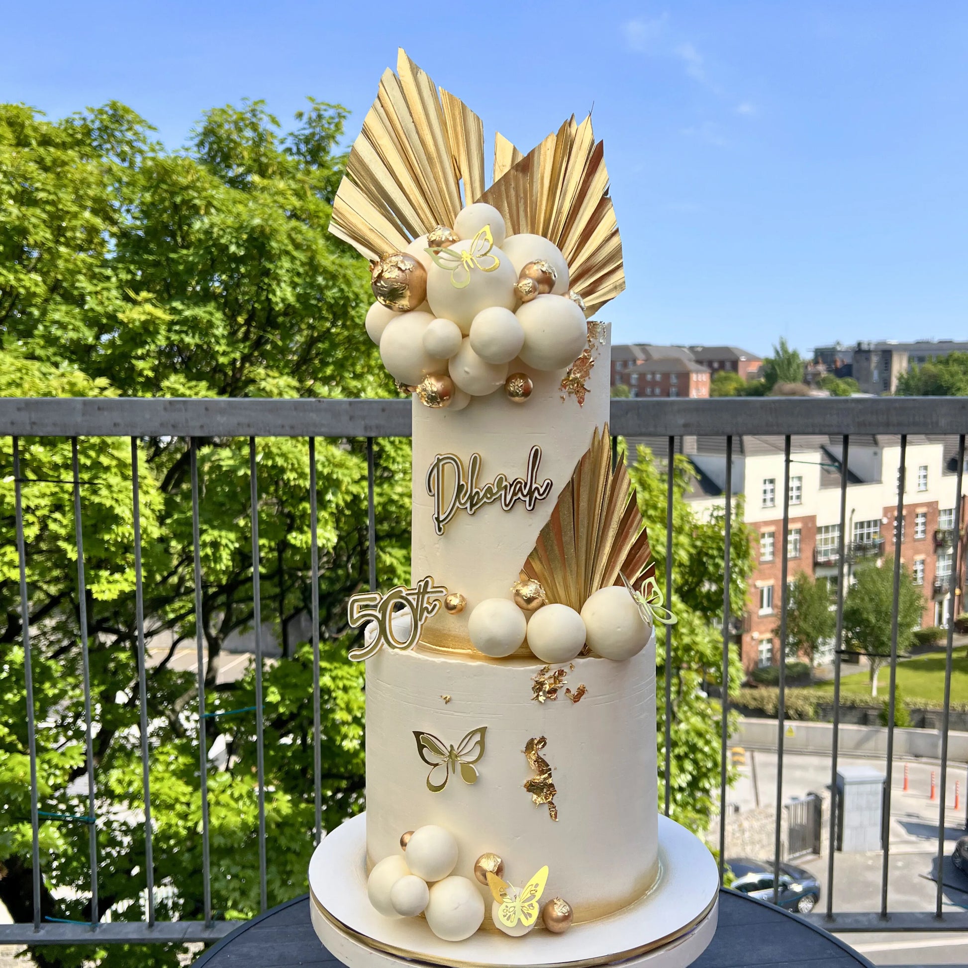 Decorative cake with gold and white elements on a balcony with greenery and buildings in the background