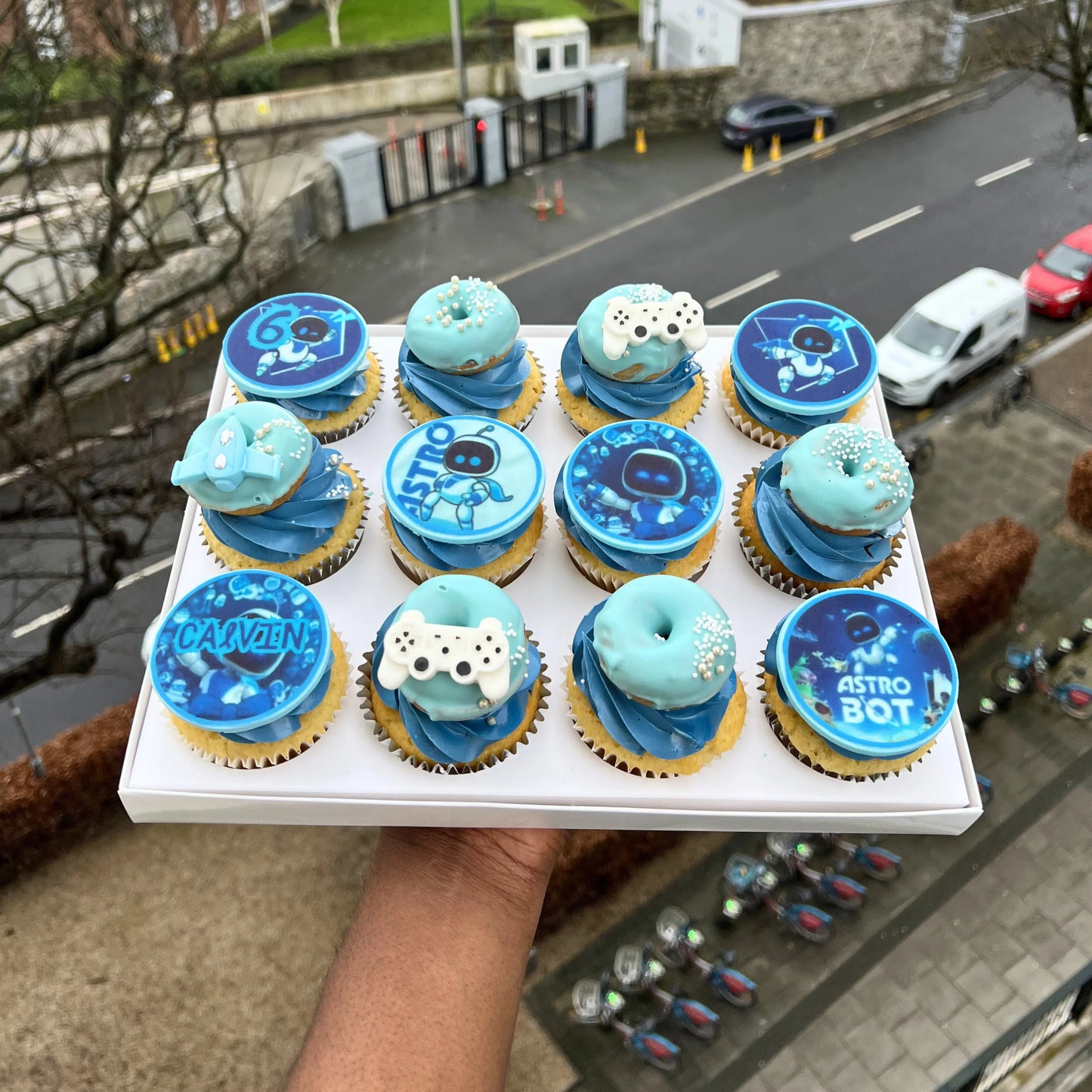 Tray of cupcakes with Astro Bot theme held by a person outdoors.