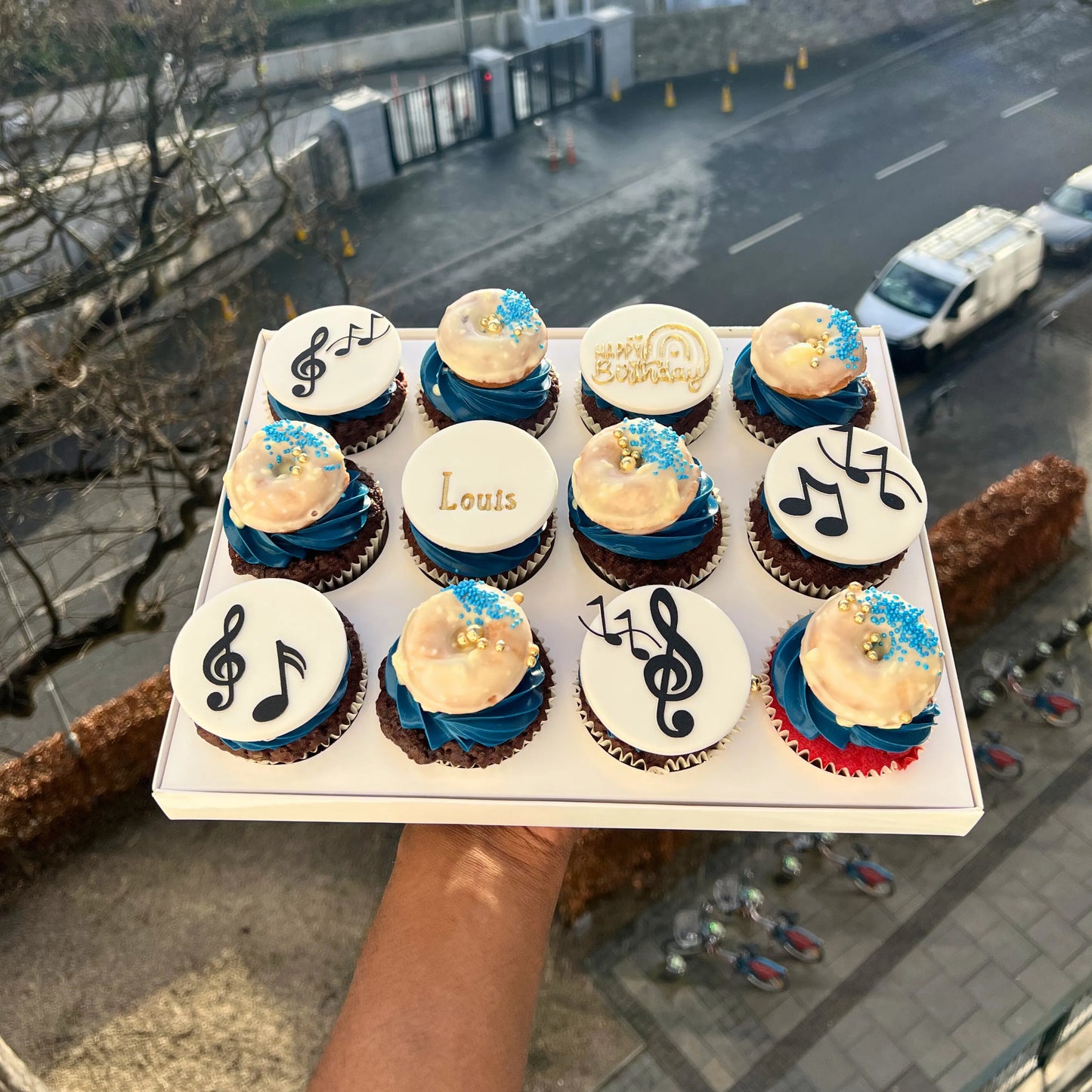 Tray of cupcakes with musical designs held by a person on a street.