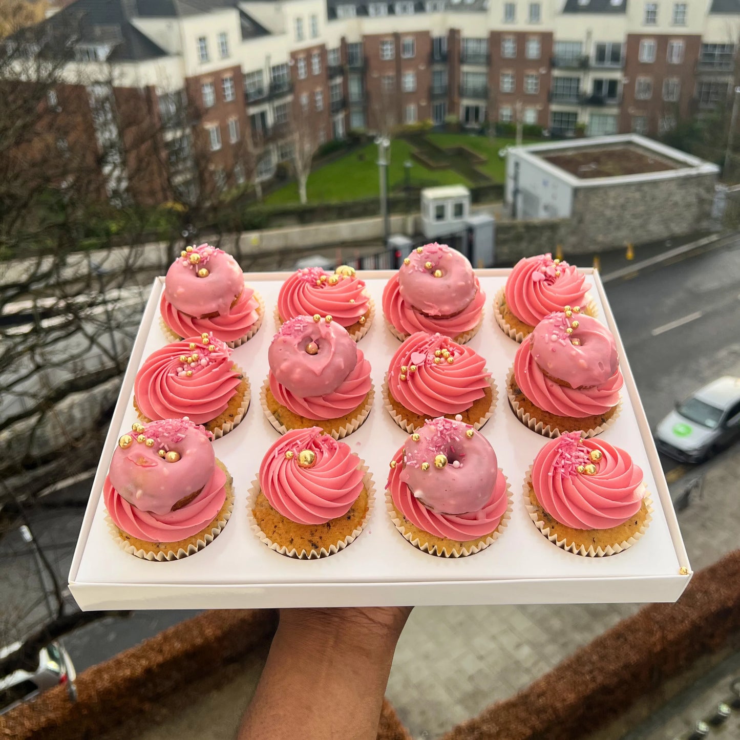 Tray of cupcakes with pink frosting held in front of a cityscape