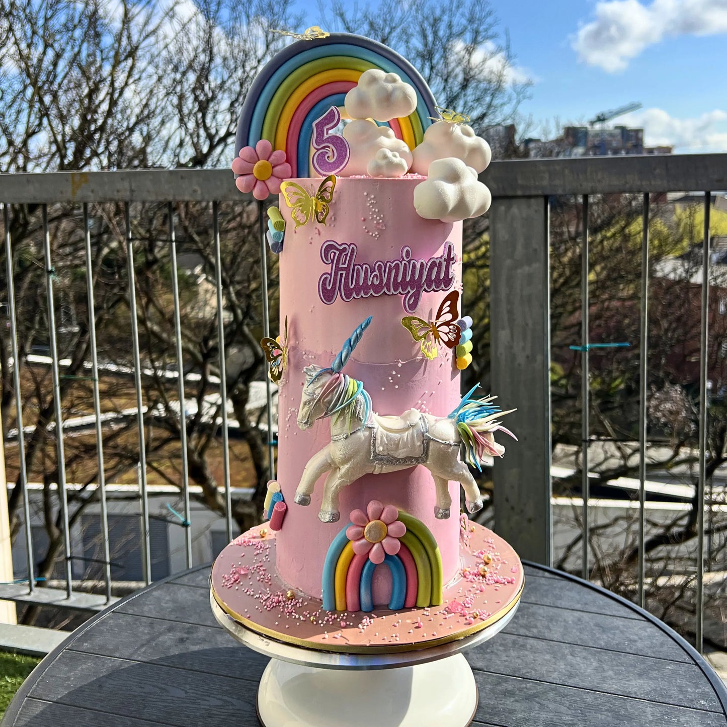 Colorful birthday cake with unicorn and rainbow decorations on a stand outdoors.