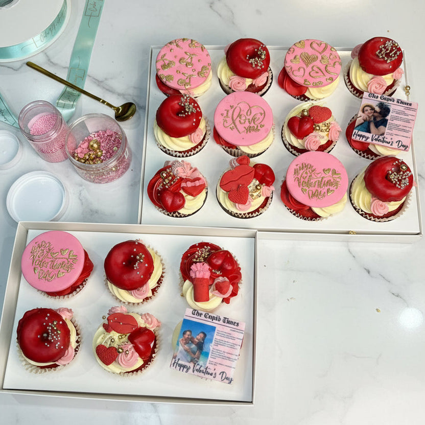 Two boxes of Valentine's Day-themed cupcakes with pink and red decorations on a gray surface.