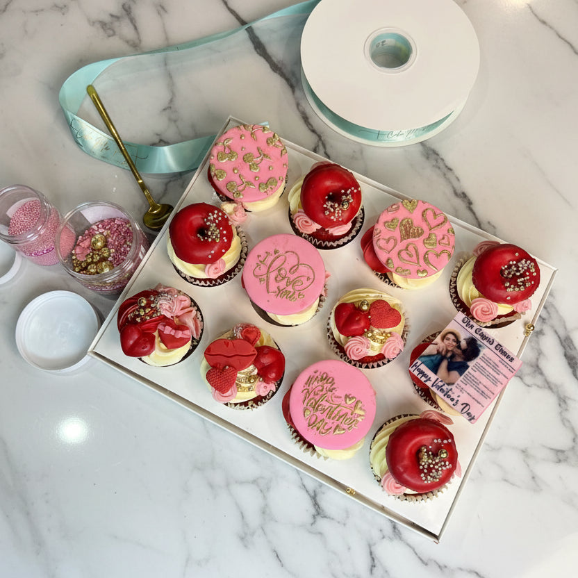 Box of decorated cupcakes with pink and red frosting on a gray surface.