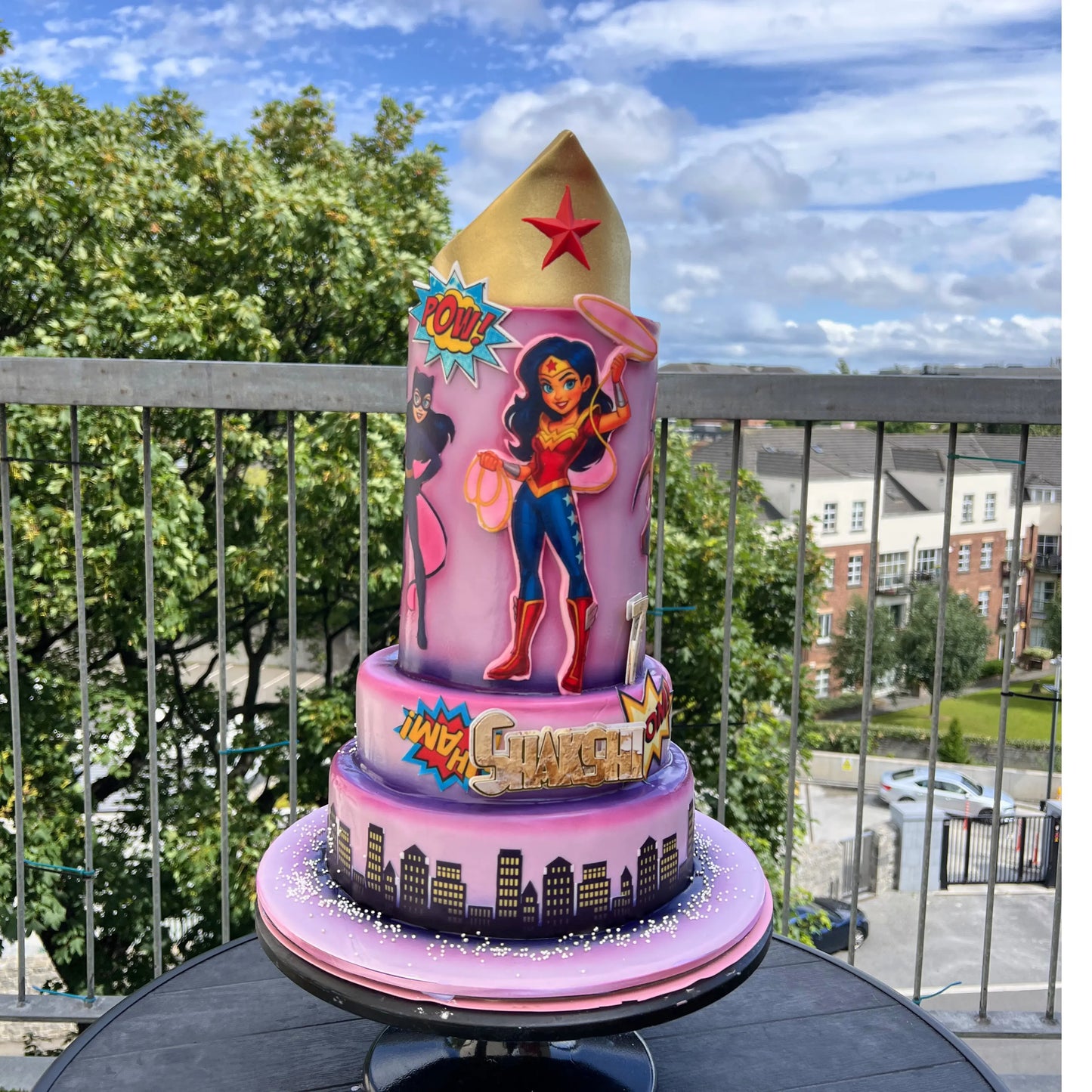 Wonder Woman-themed cake on a balcony with trees and buildings in the background