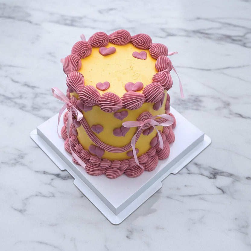Small yellow cake with pink decorative elements on a white tiled floor.
