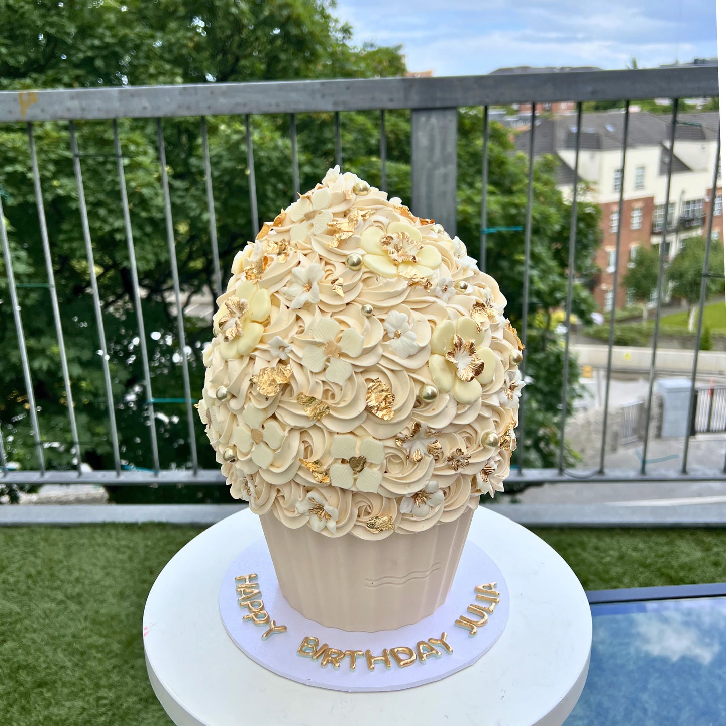 Large decorative cupcake with white frosting and gold accents on a patio with greenery and buildings in the background.