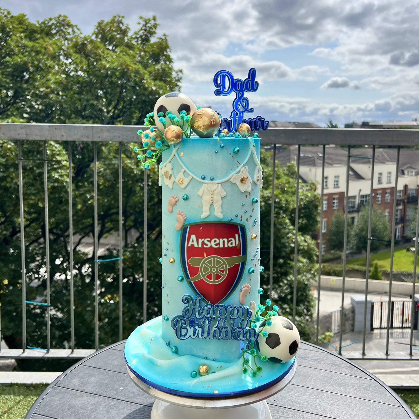 Decorative cake with Arsenal logo and 'Dad & Son' topper on a balcony with trees and buildings in the background