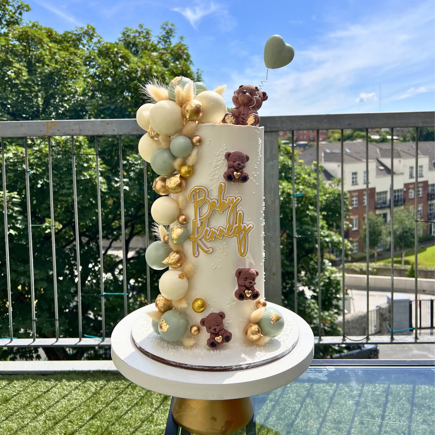 beige christening cake with teddy bears and balloons on a balcony with a view of trees and buildings.
