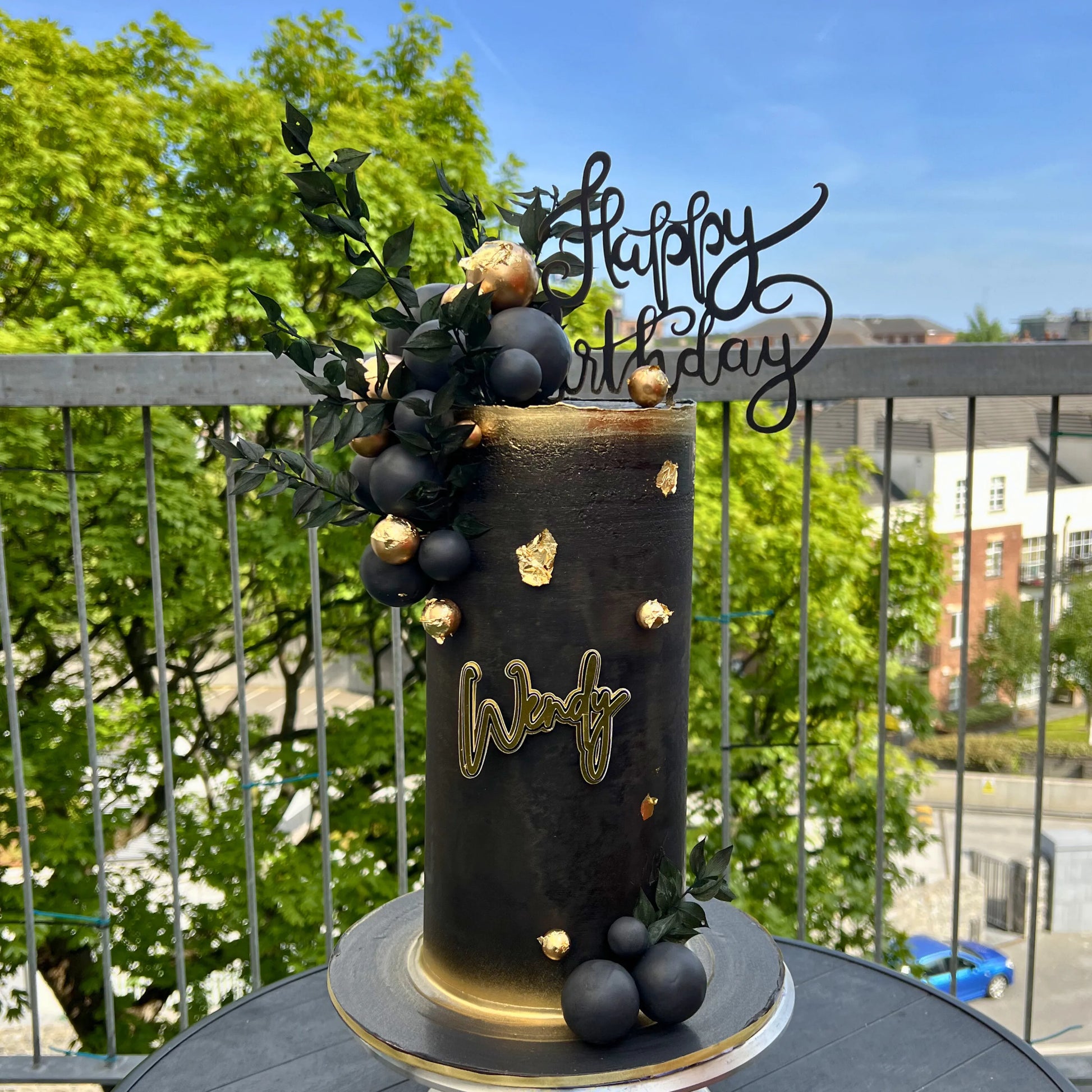 Decorative birthday cake with 'Happy Birthday' topper on a balcony with greenery and buildings in the background