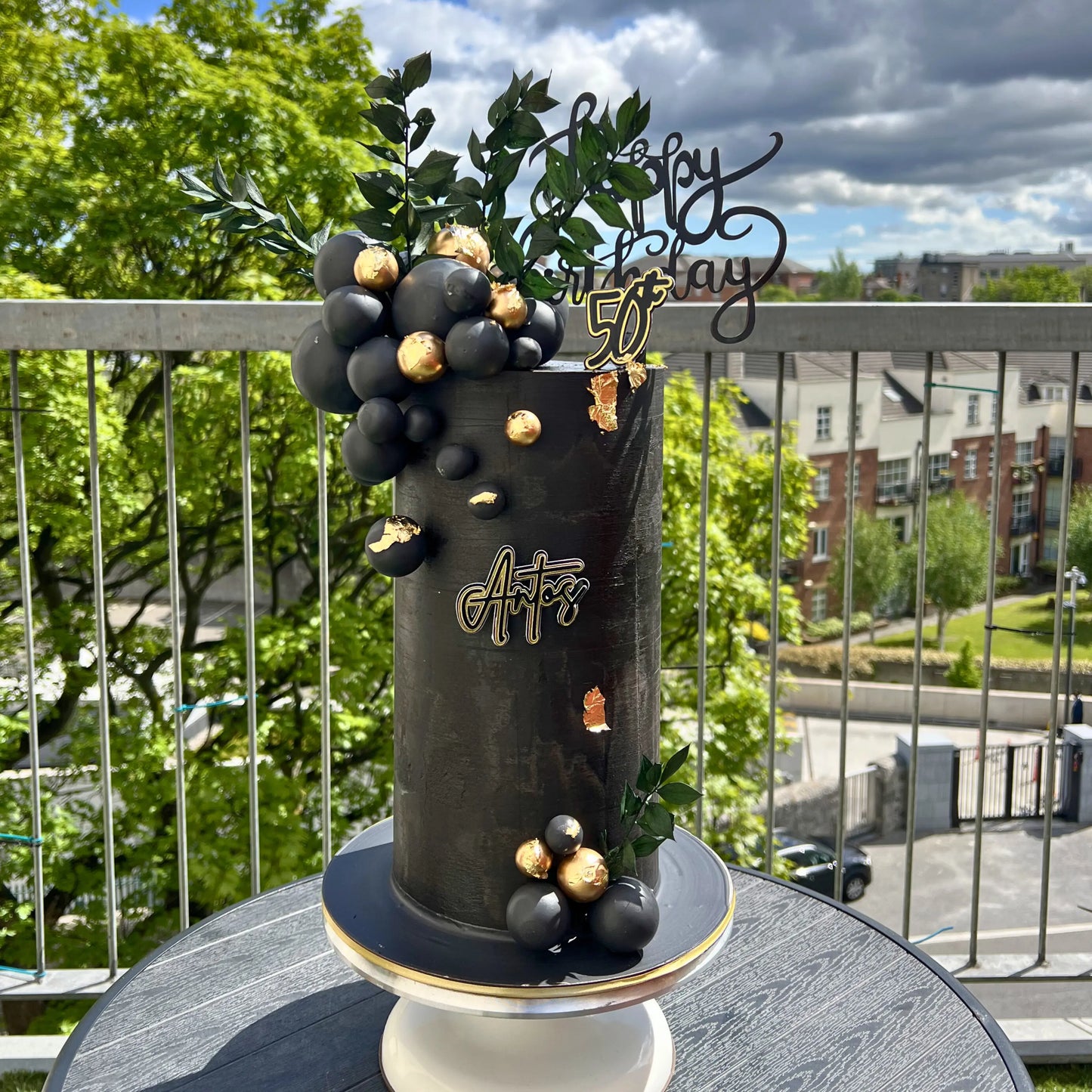 Decorative cake with black and gold grapes on a balcony with greenery and buildings in the background
