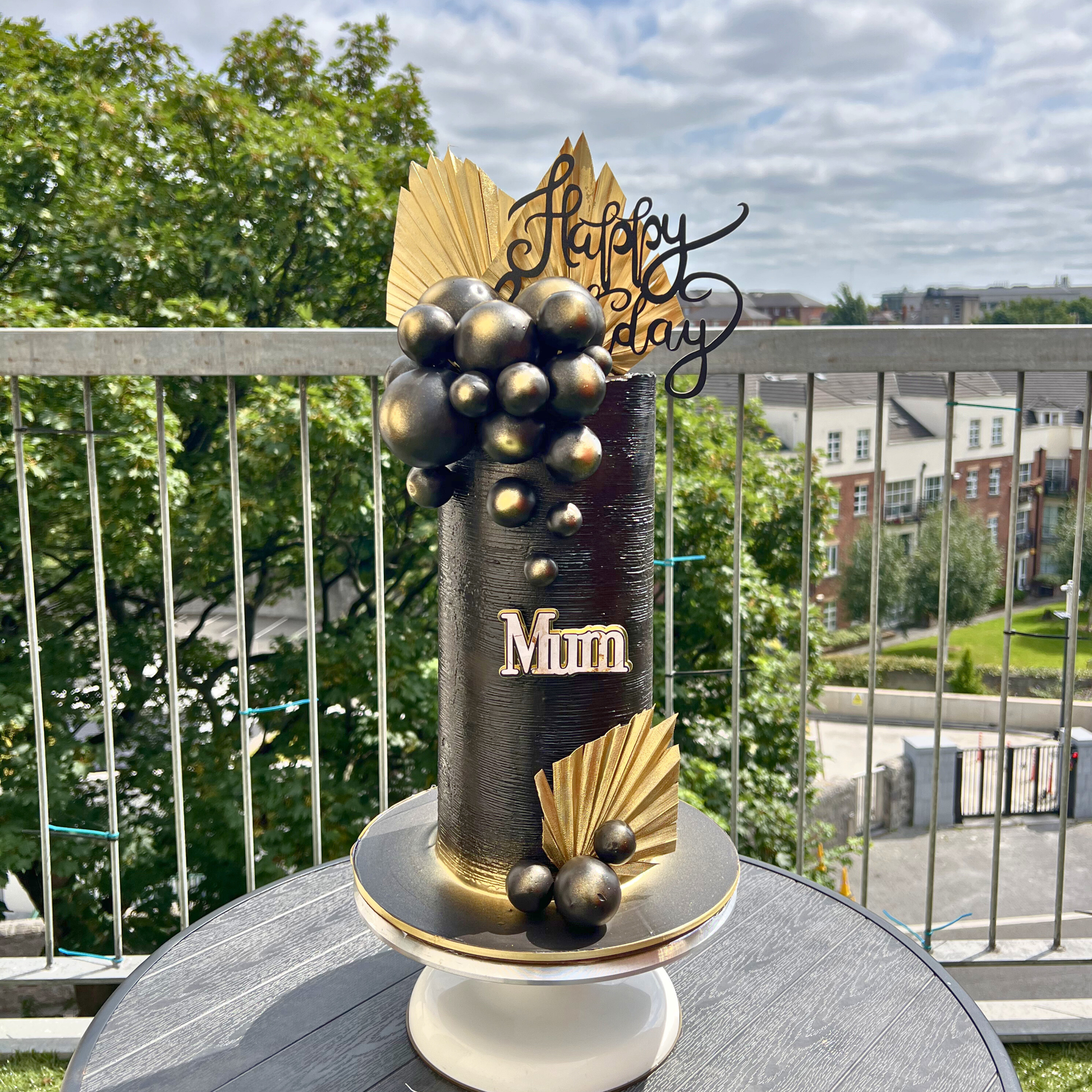 Decorative cake with 'Happy BirthDay' and 'Mum' text on a balcony with greenery and buildings in the background.
