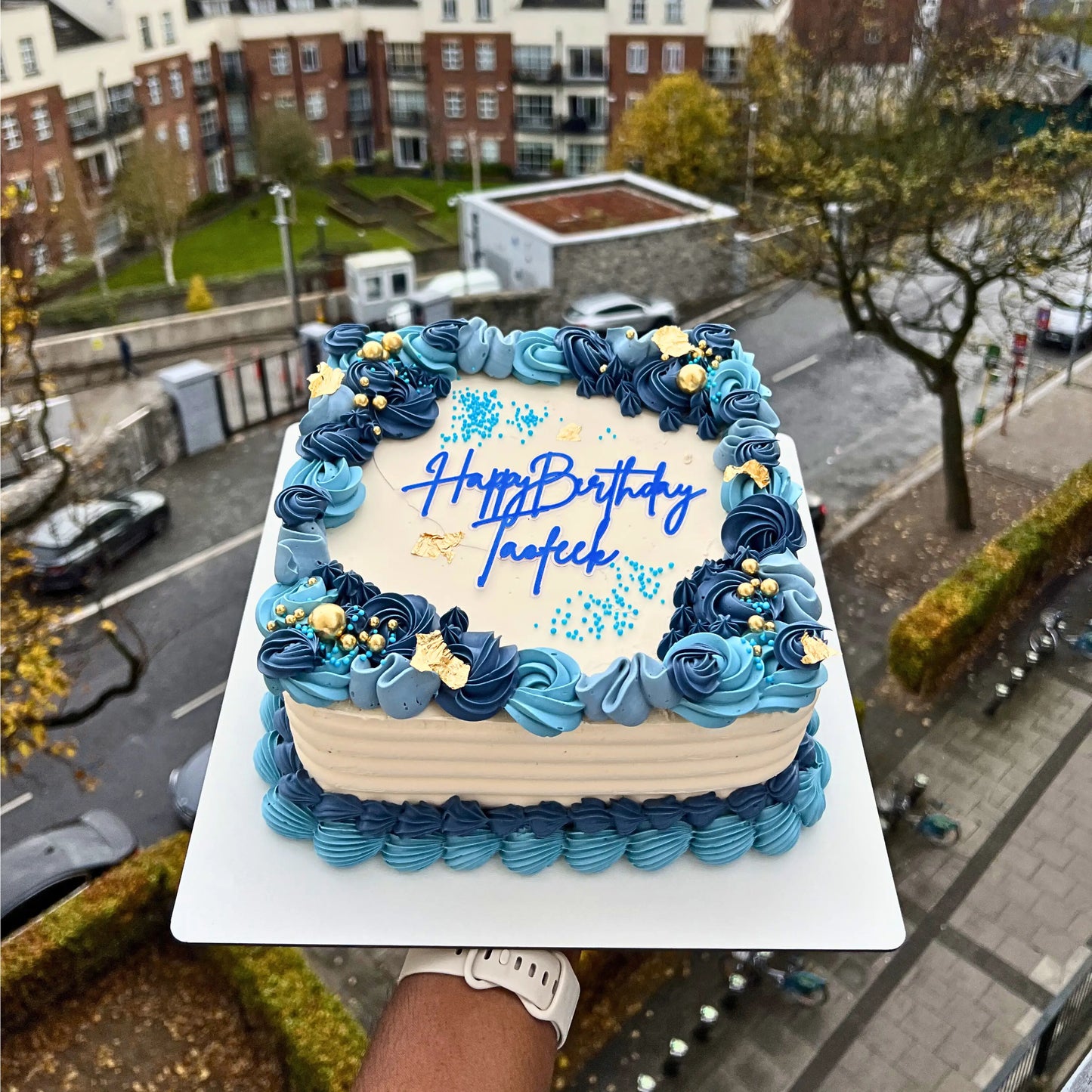 Rectangular birthday cake with blue and beige frosting, placed on a street with buildings and cars in the background.