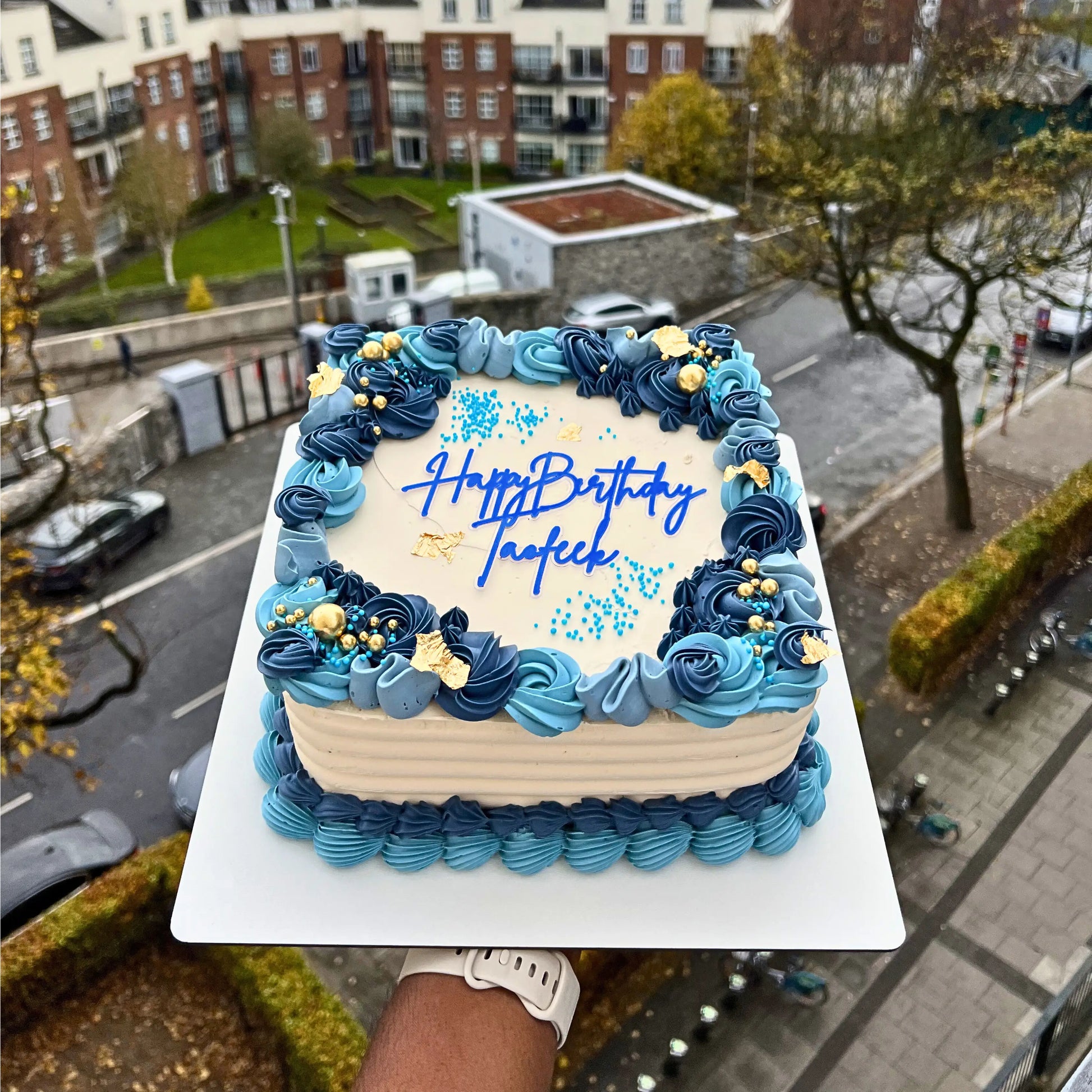 Rectangular birthday cake with blue and beige frosting, placed on a street with buildings and cars in the background.