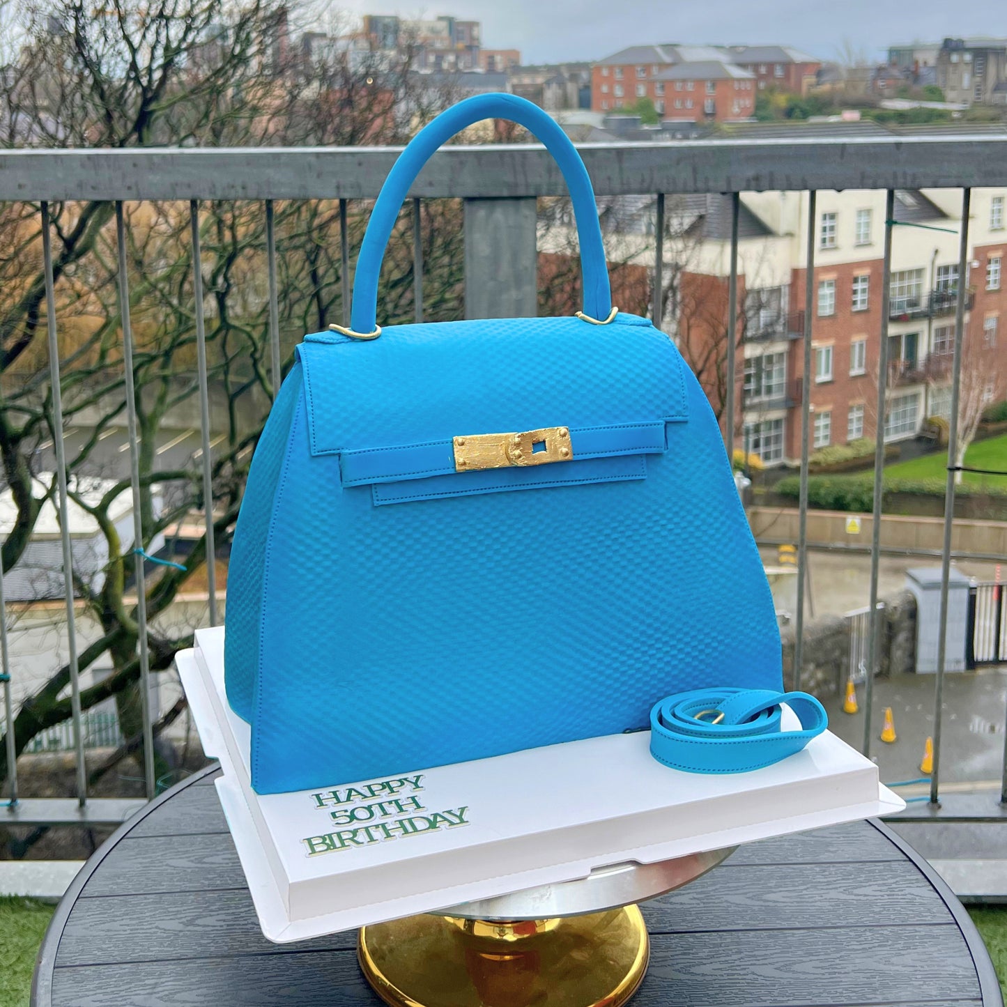 A bright blue textured handbag-shaped cake displayed on a white book with 'Happy 50th Birthday' printed on it, set on a round table outdoors with a balcony railing and residential buildings in the background.