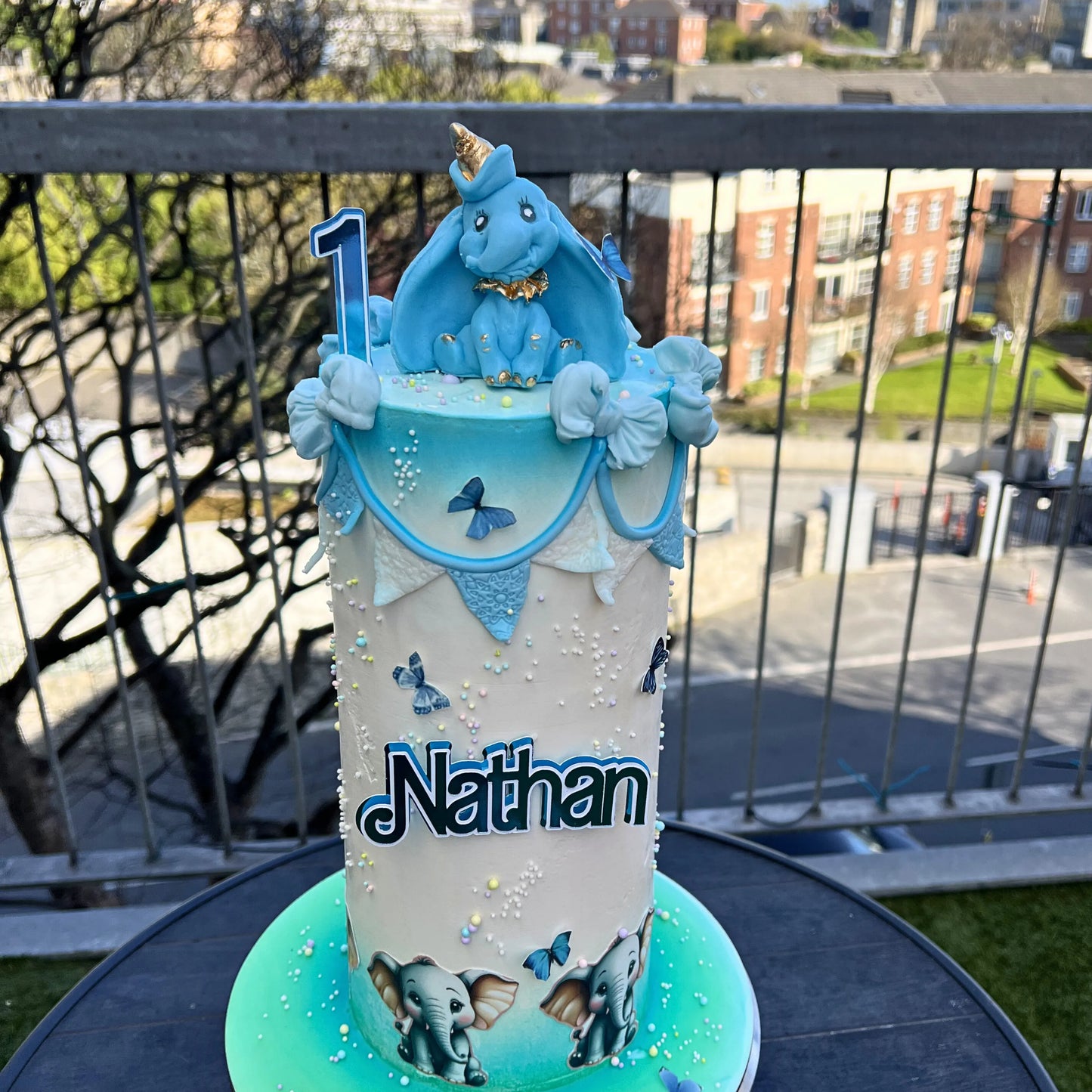 Decorative cake with blue and white elephant theme, featuring the name 'Nathan', on a balcony with a cityscape background.