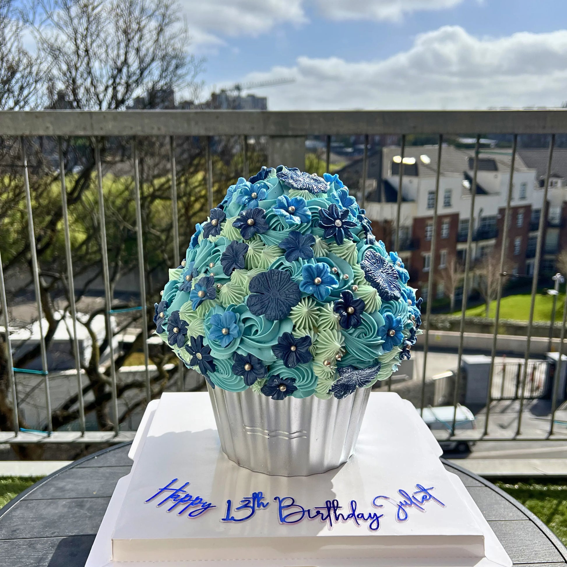 Decorative cake with blue and green flowers on a silver cupcake base, placed on a white card with 'Happy 13th Birthday' text, outdoors.