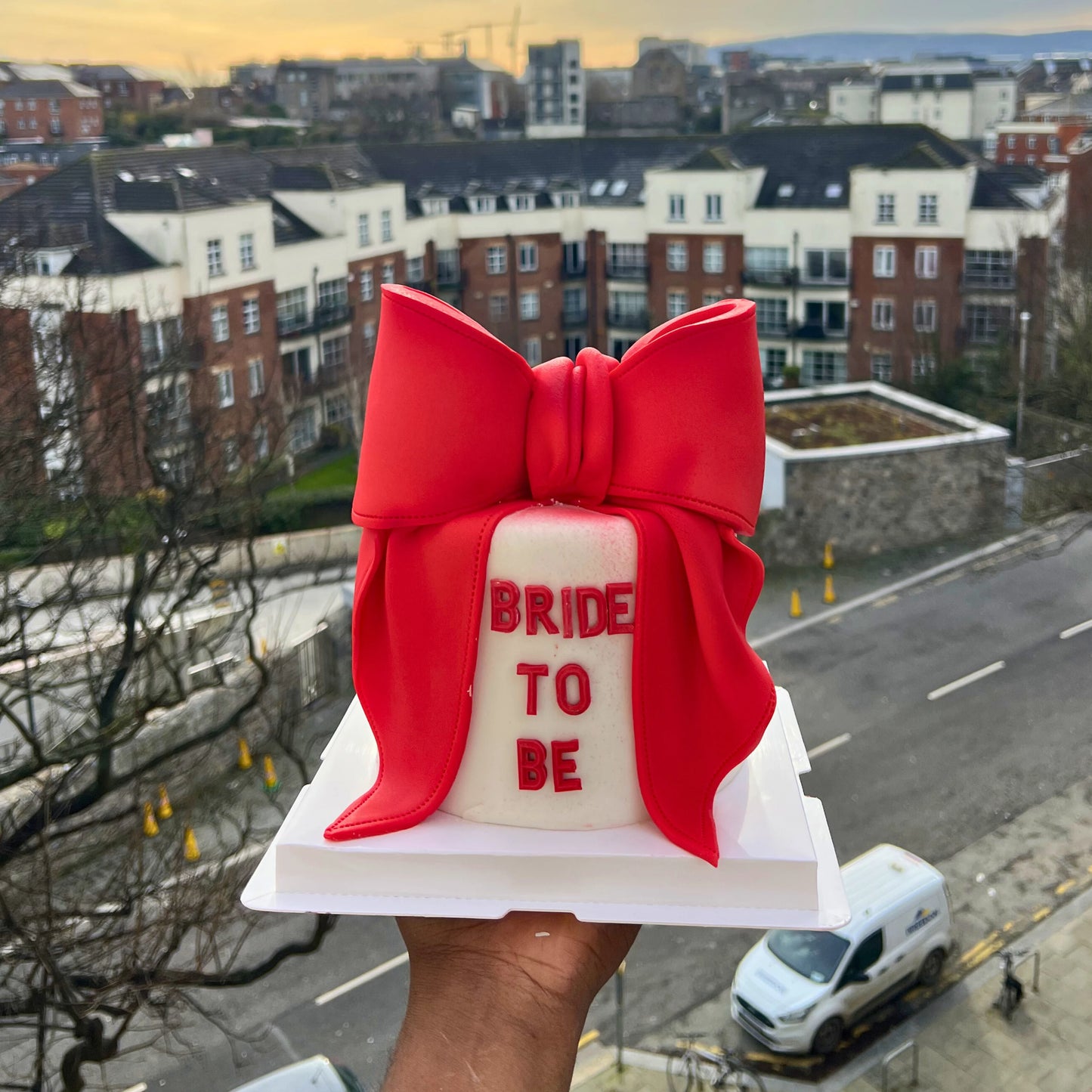 Cake with a red bow and 'Bride to Be' text held up against a cityscape background
