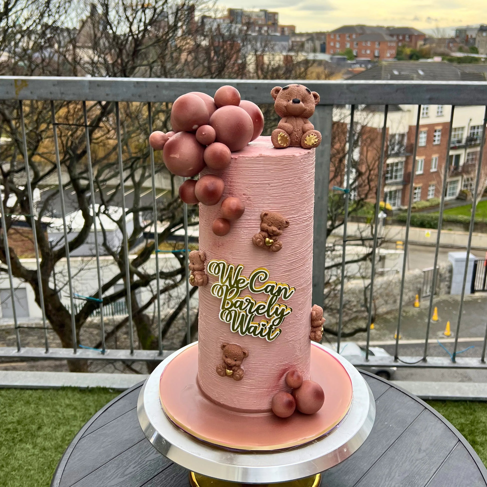 Pink cake with teddy bear decorations and 'We Can Barely Wait' text, outdoors with a cityscape background.