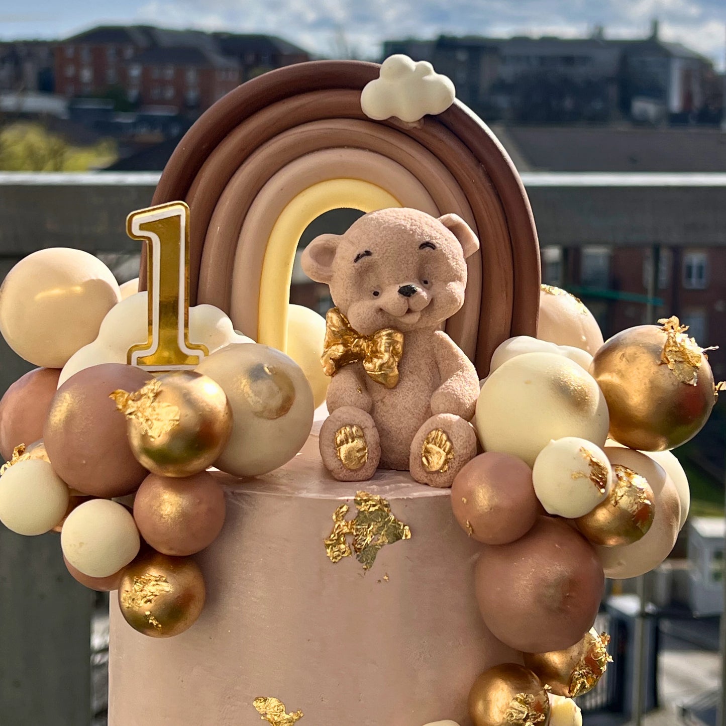 Birthday cake with teddy bear and rainbow decoration against a cityscape background