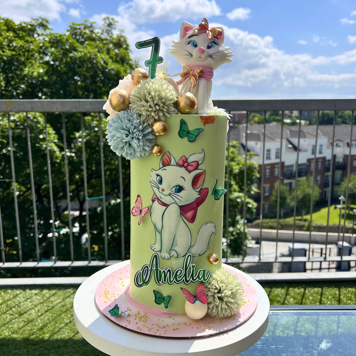 Decorative cake with cartoon cat design on a balcony with greenery and buildings in the background