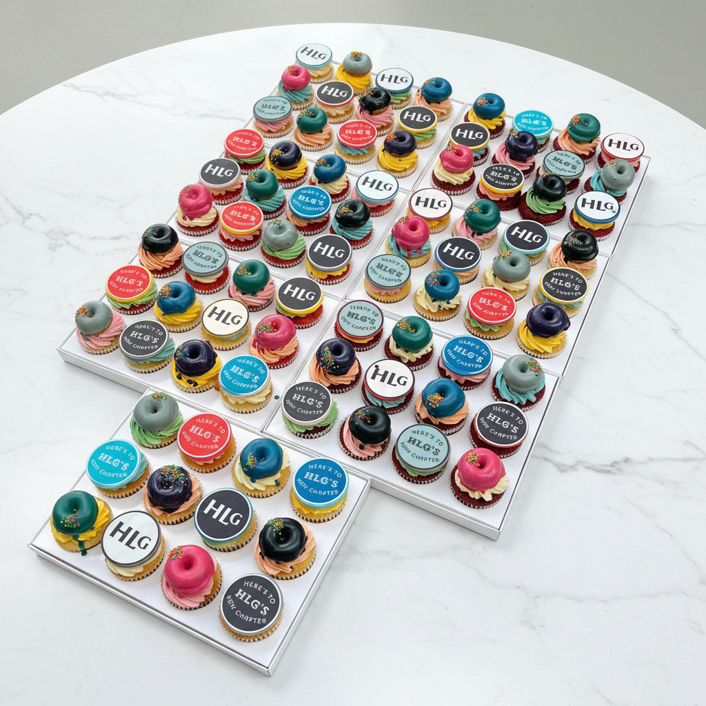 Multiple white cupcake trays filled with assorted frosted cupcakes arranged on artificial grass. Each cupcake has a round branded topper reading 'HLG' in white lettering, and the cupcakes feature colorful frosting in shades of blue, pink, teal, purple, red, and black.