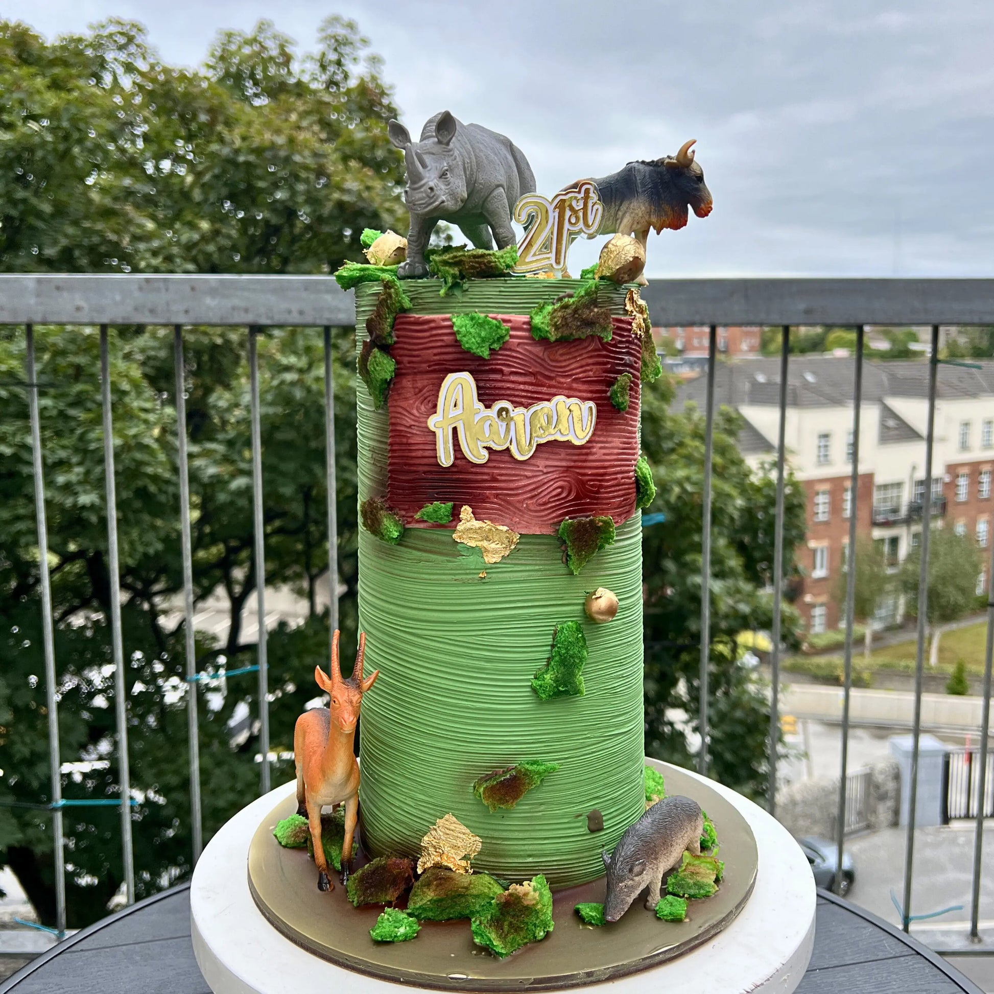 Decorative cake with animal figures and 'Aeran' branding on a balcony with trees and buildings in the background.