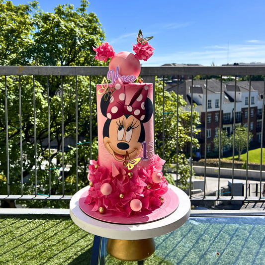 Pink Minnie Mouse-themed birthday cake on a balcony with trees and buildings in the background