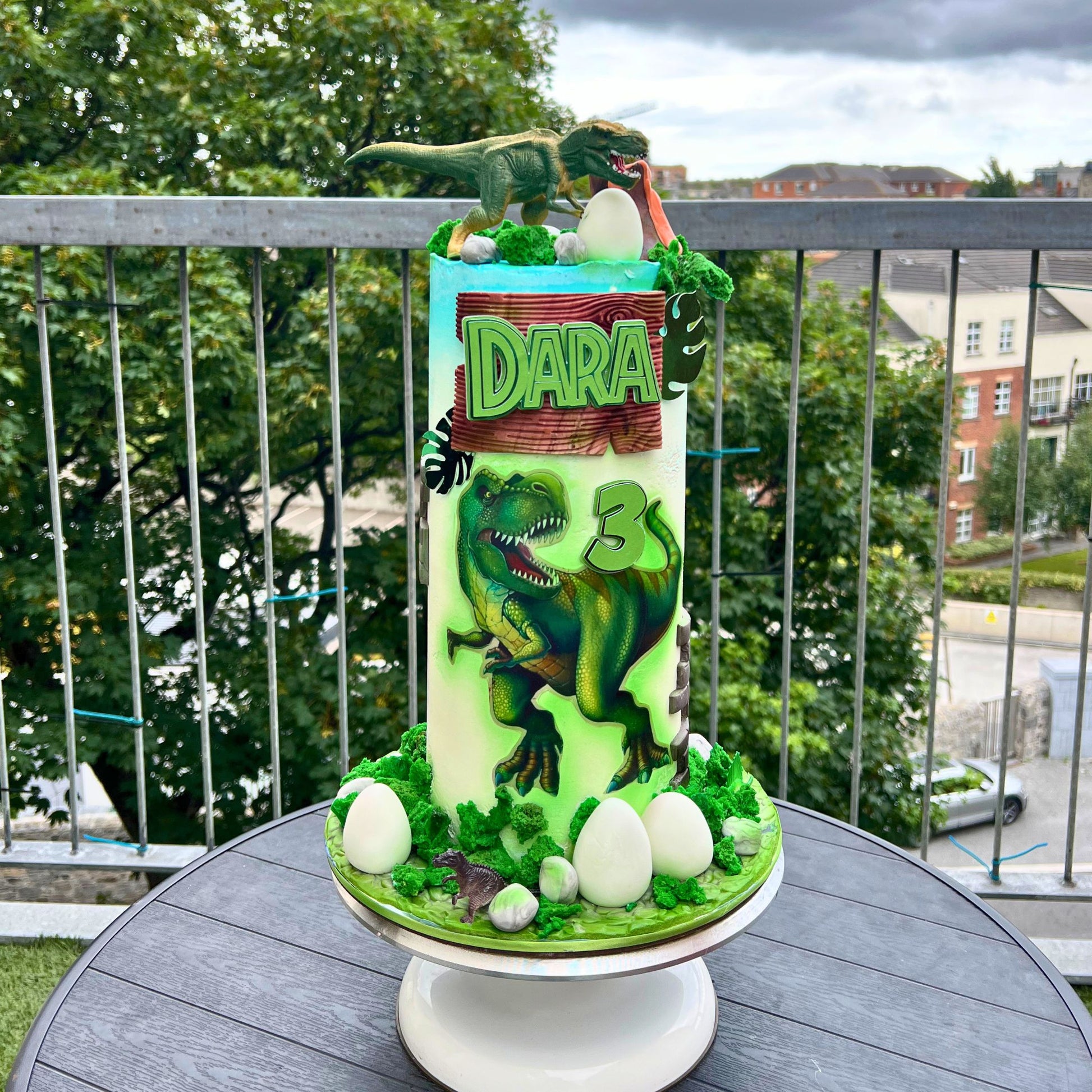 A custom dinosaur-themed birthday cake on a white pedestal stand, set on a dark gray outdoor table with a balcony railing and trees in the background. The cake is decorated with green icing, a large green T-Rex, white egg toppers, and a wooden sign reading 'DARA'.